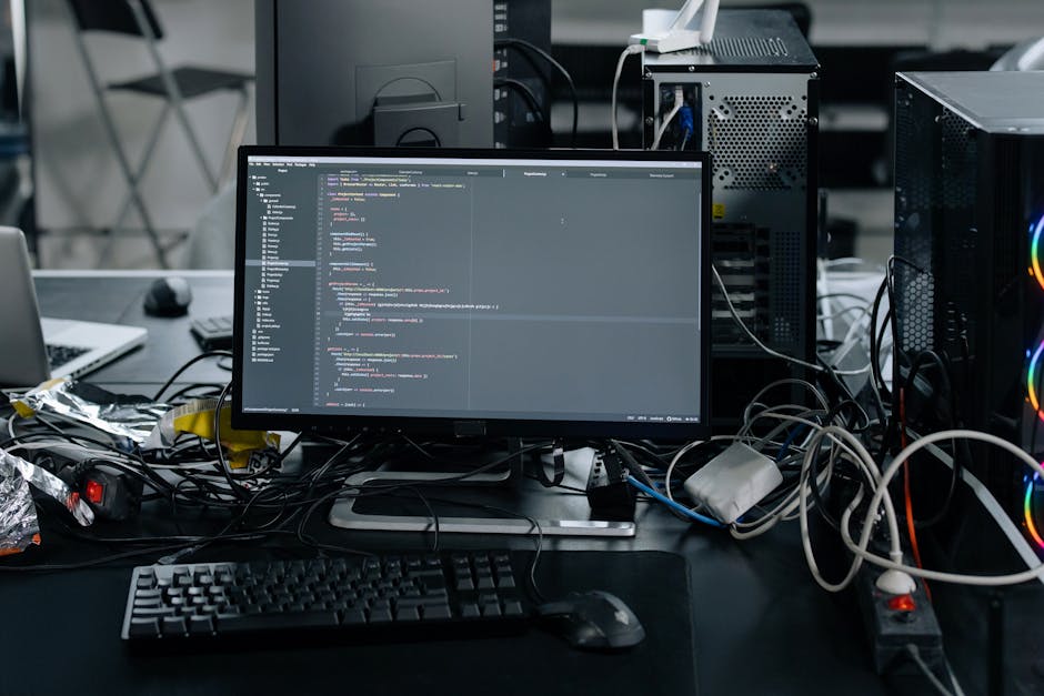 A computer monitor displaying lines of programming code on a cluttered desk surrounded by tangled cables, a keyboard, a mouse, and electronic devices, in a workspace environment that appears disorgani